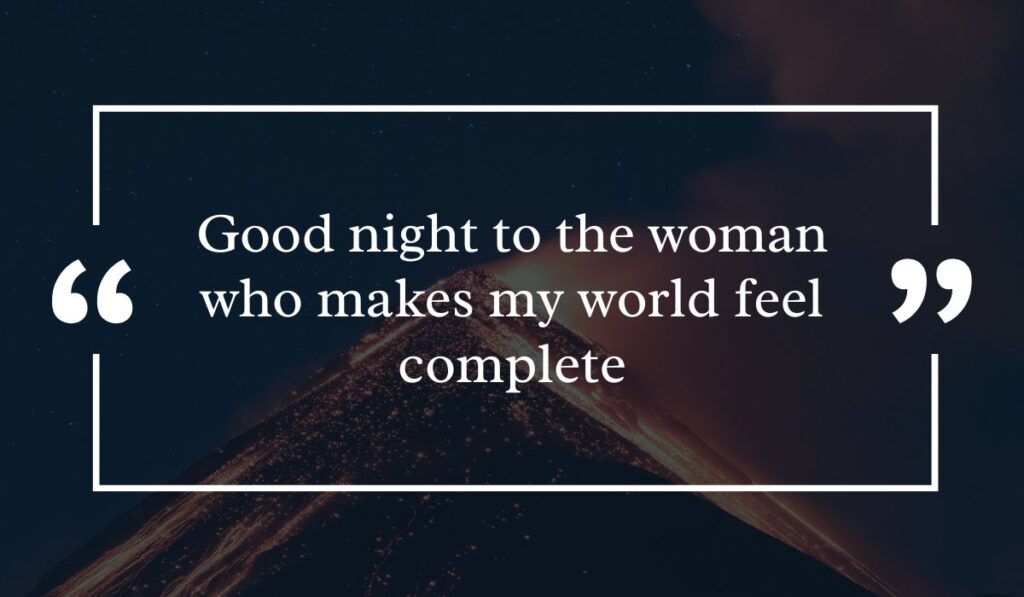 Dramatic dark night sky background featuring a lit volcanic peak at night, centered with a white quotation box highlighting heartfelt good night text messages for her: "Good night to the woman who makes my world feel complete".