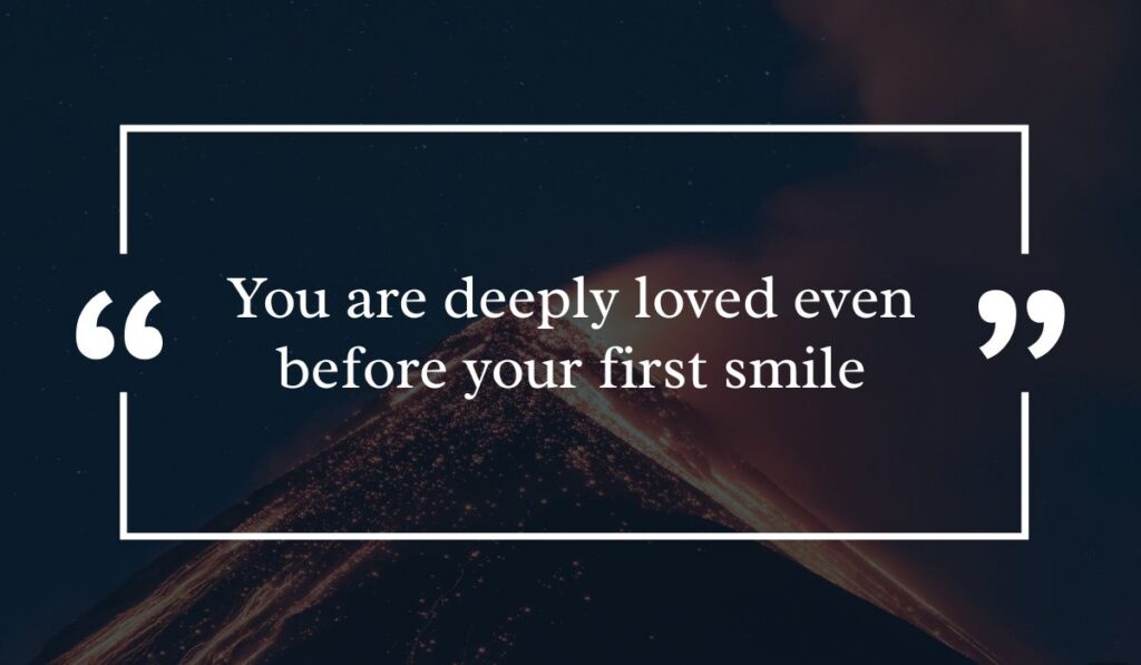 Dramatic dark night sky background featuring a lit volcanic peak at night, centered with a white quotation box highlighting touching wishes for new born baby girl: "You are deeply loved even before your first smile".